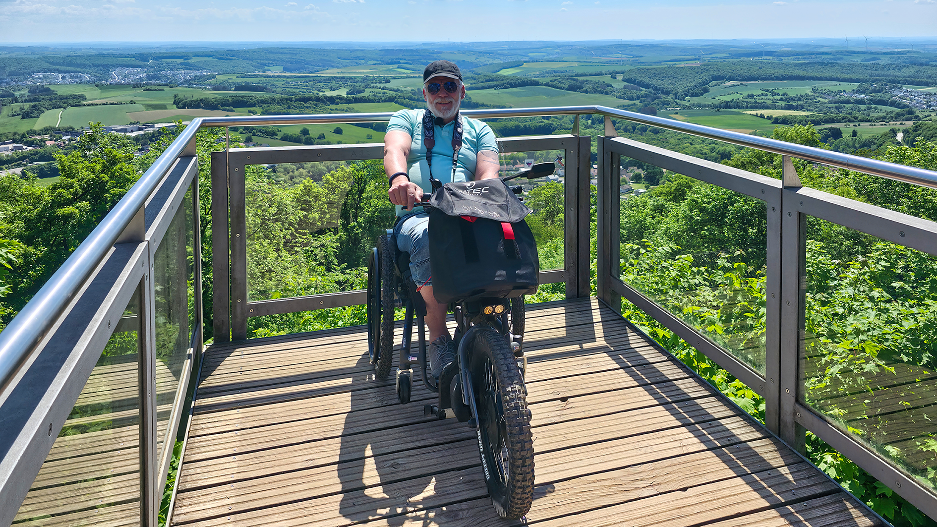 Das Bild zeigt Hans-Jürgen Rohe auf einer Aussichtsplattform über dem Saarland mit seinem Rollstuhl SCRAMBLER 2 und dem Rollstuhlzuggerät von BATEC und AAT.
